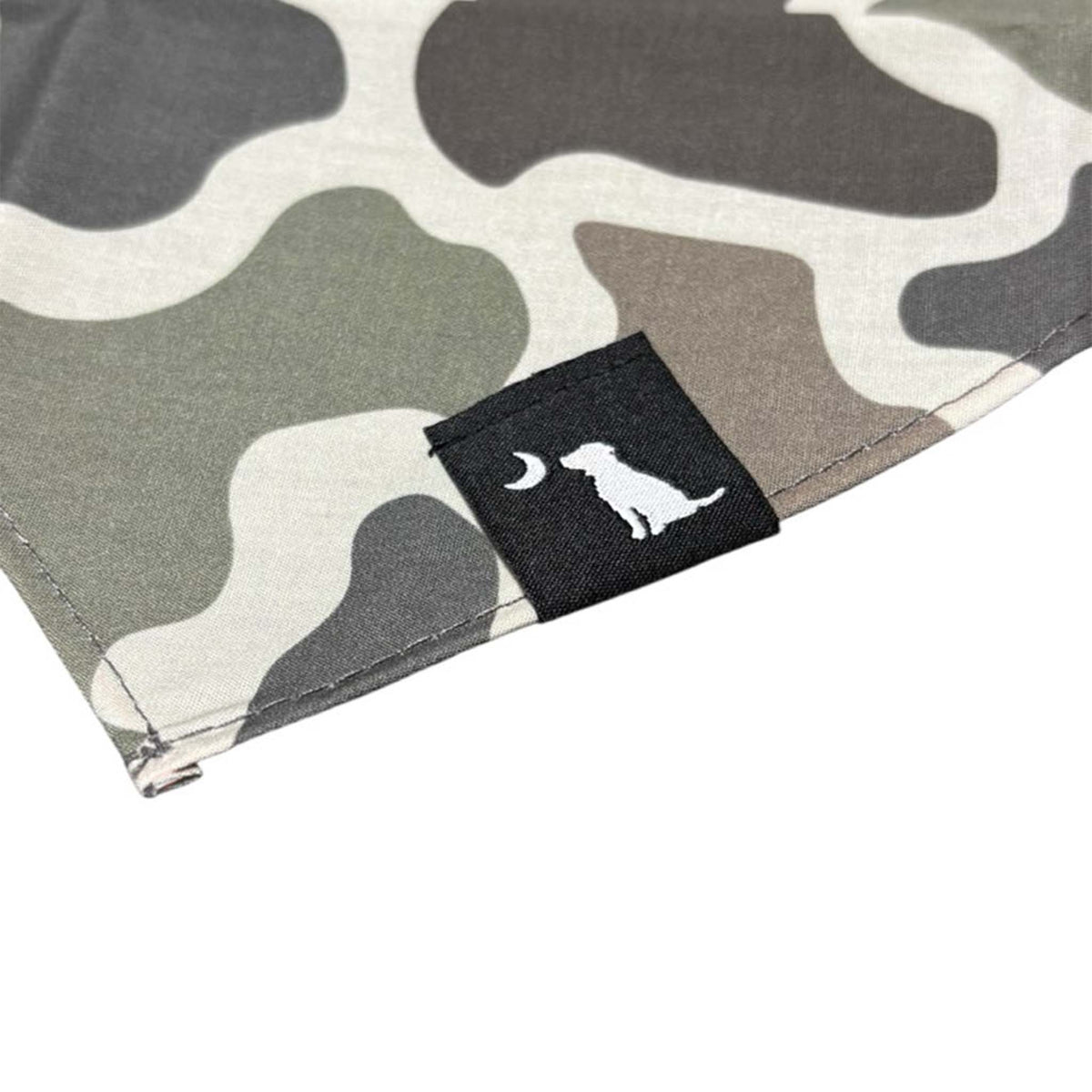 Close-up of camo print dog bandana in tan, olive, and brown with black fabric tag featuring a white dog and crescent moon.