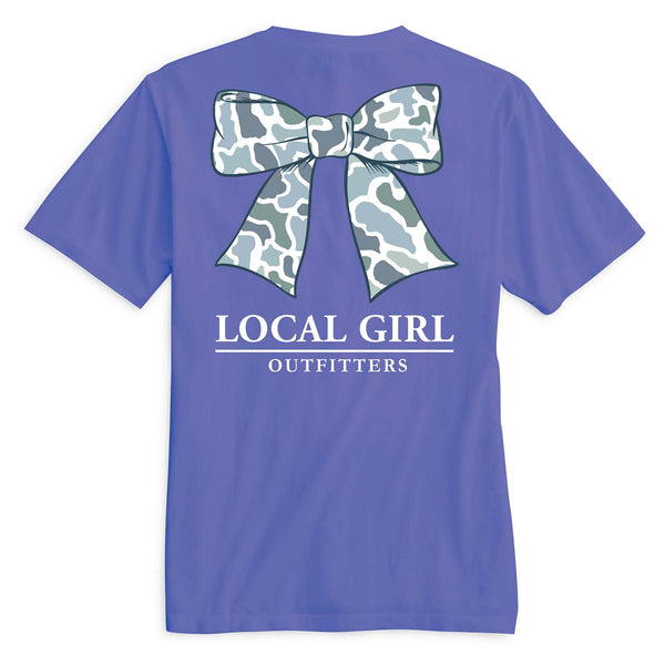 Back view of a violet t-shirt featuring a large bow graphic with a gray and white camo pattern. Below the bow, the text "LOCAL GIRL" is printed in white uppercase letters, with the word "OUTFITTERS" underneath separated by a horizontal line.