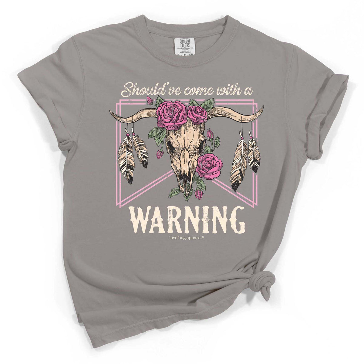 Grey short sleeve t-shirt with graphic of a steer skull adorned with pink roses and feathers, framed by pink lines, and the text “Should’ve come with a WARNING” across the front.