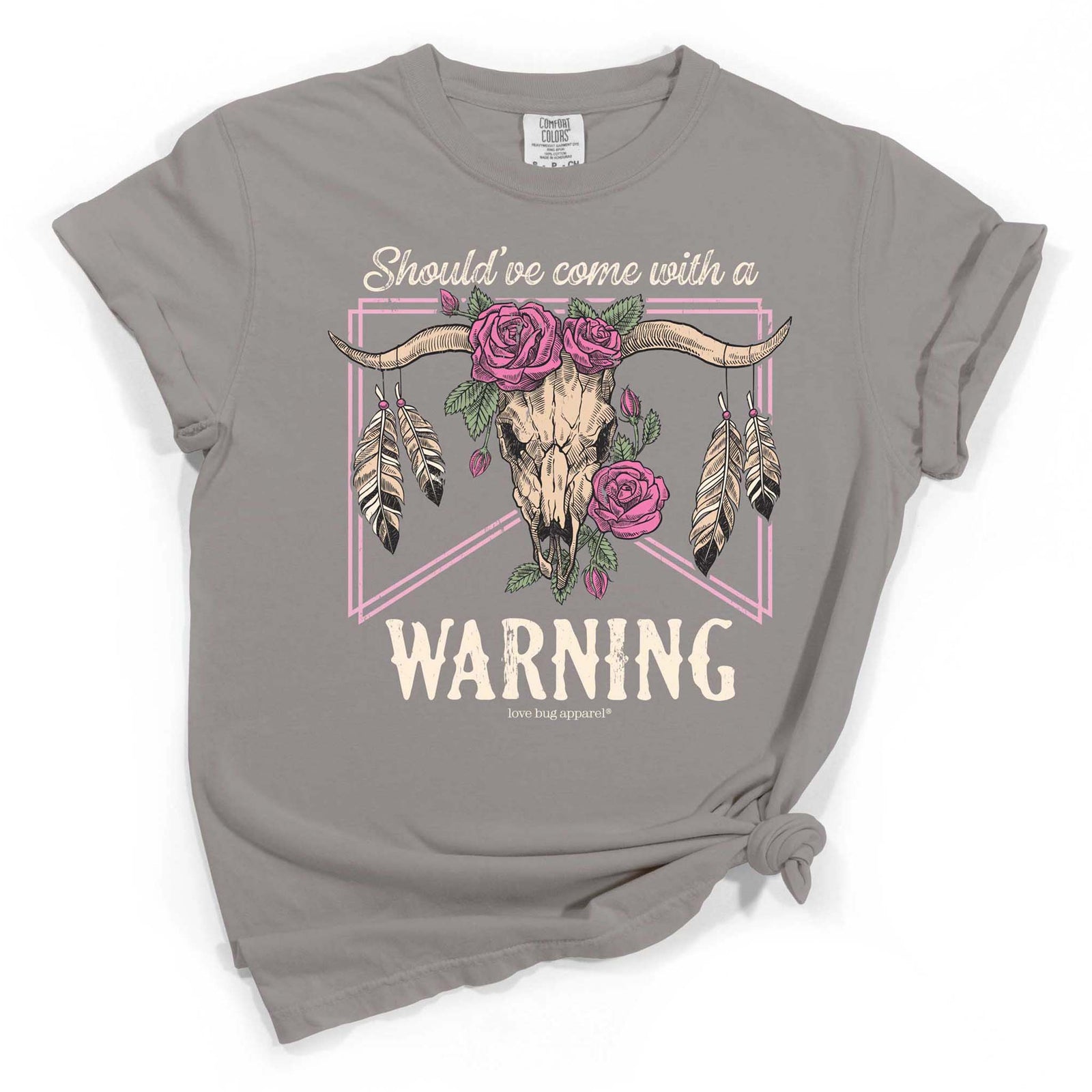 Grey short sleeve t-shirt with graphic of a steer skull adorned with pink roses and feathers, framed by pink lines, and the text “Should’ve come with a WARNING” across the front.