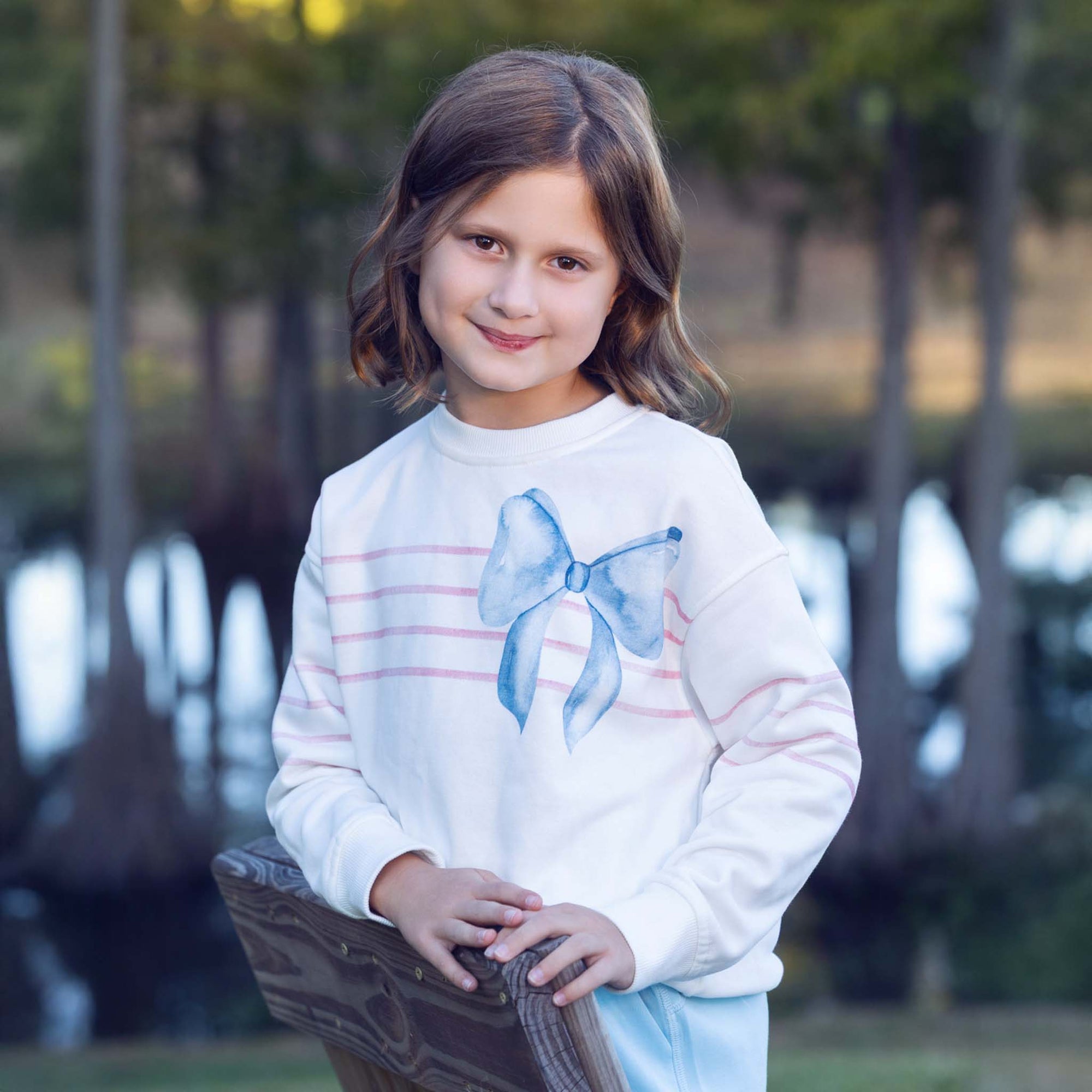 Young girl wearing a white Love Bug sweatshirt featuring a blue bow graphic and pink stripes across the chest, standing outdoors near a lake and trees.