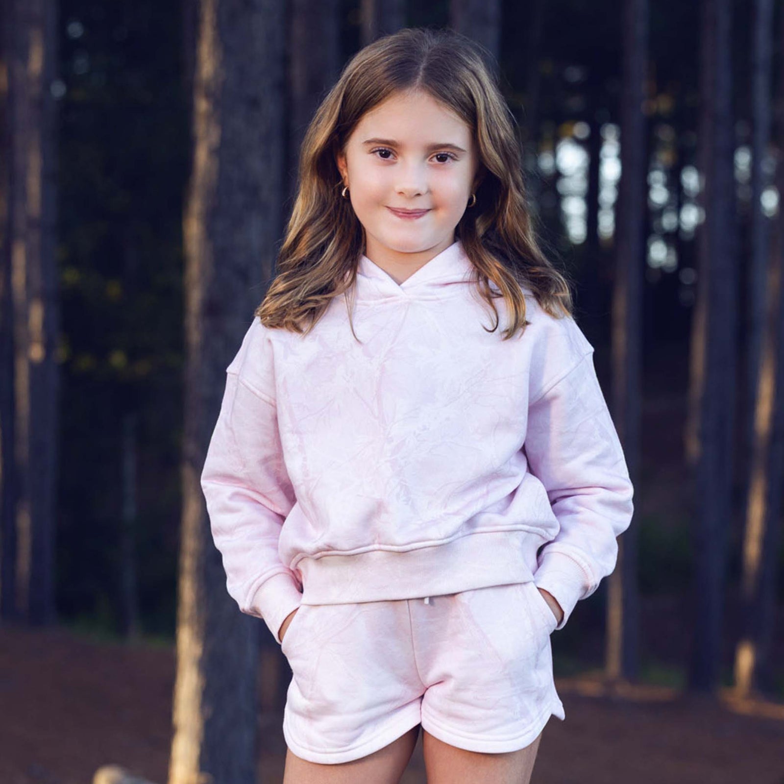 Young girl wearing a pink hoodie and shorts set standing in front of a dark background.