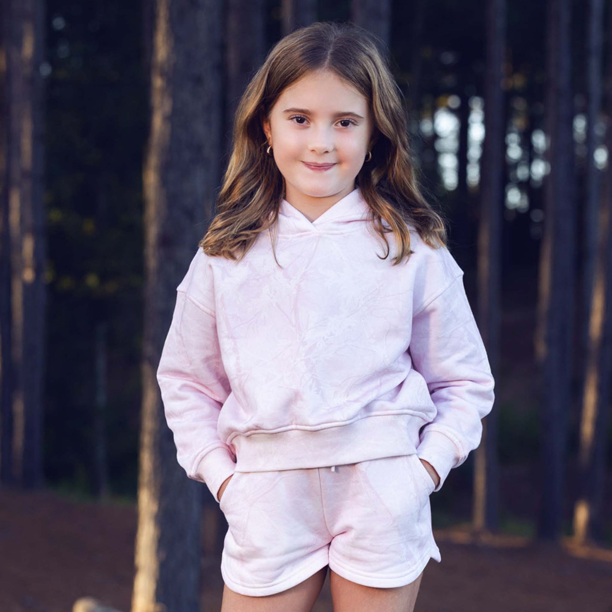 Young girl wearing a pink hoodie and shorts set standing in front of a dark background.