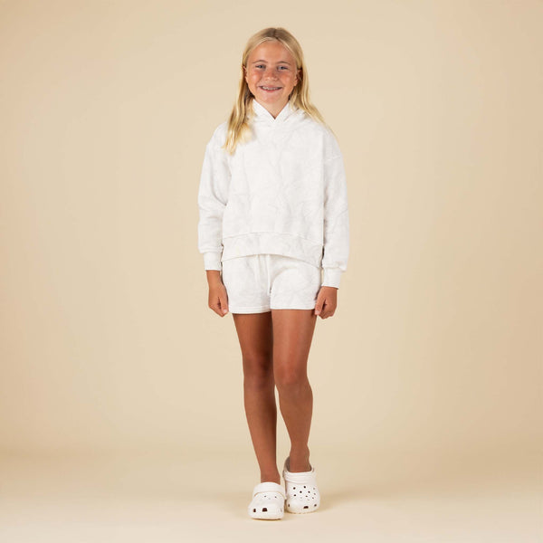 Full-body front view of girl in white tonal camo hoodie and shorts with white Crocs, standing against beige background.