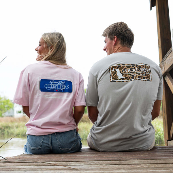 A woman and man sit side by side on a wooden dock, facing away from the camera and smiling at each other. The woman wears a light pink t-shirt with a blue and white “Local Girl Outfitters” logo on the back, paired with denim shorts. The man wears a light gray t-shirt with a camo-print “Local Boy Outfitters” logo on the back. The background features marsh grass and calm water under a cloudy sky.