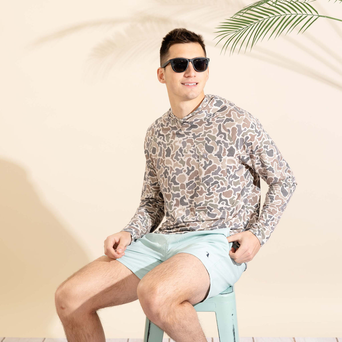 Seated male model wearing a camo long sleeve shirt and light blue shorts against a beige backdrop with palm leaf accents.