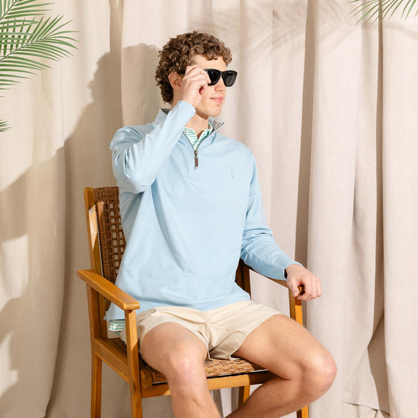 Seated male model wearing sunglasses and a light blue quarter-zip pullover with beige shorts in a neutral indoor setting.