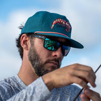 Teal snapback hat with embroidered shrimp graphic and pink rope detail worn by man fishing, paired with polarized sunglasses.