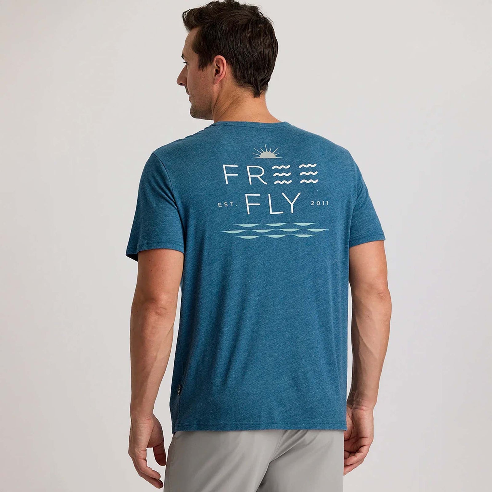 Back view of a man wearing a blue Free Fly short sleeve pocket t-shirt with a large graphic on the back. Graphic features sun, waves, and "Free Fly" text.