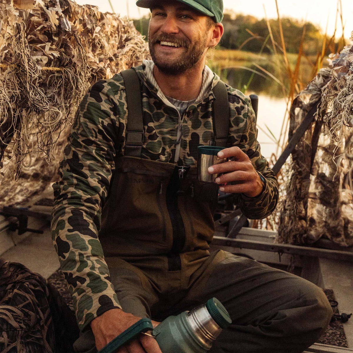 Man in camouflage clothing holding a thermos by a body of water