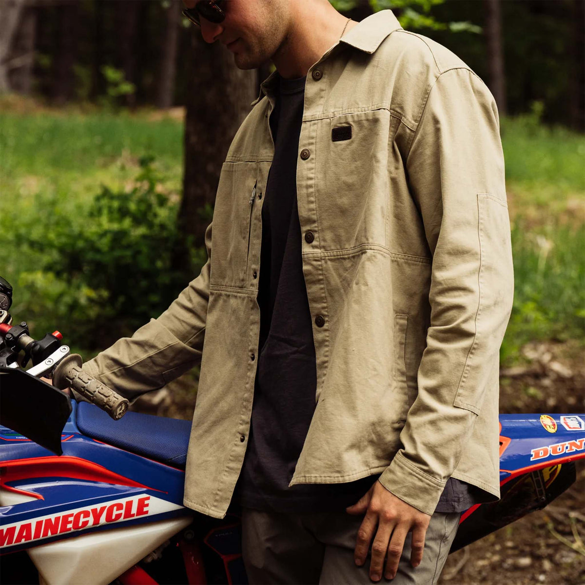 Man wearing a slate green jacket standing next to a motorcycle in a forest setting