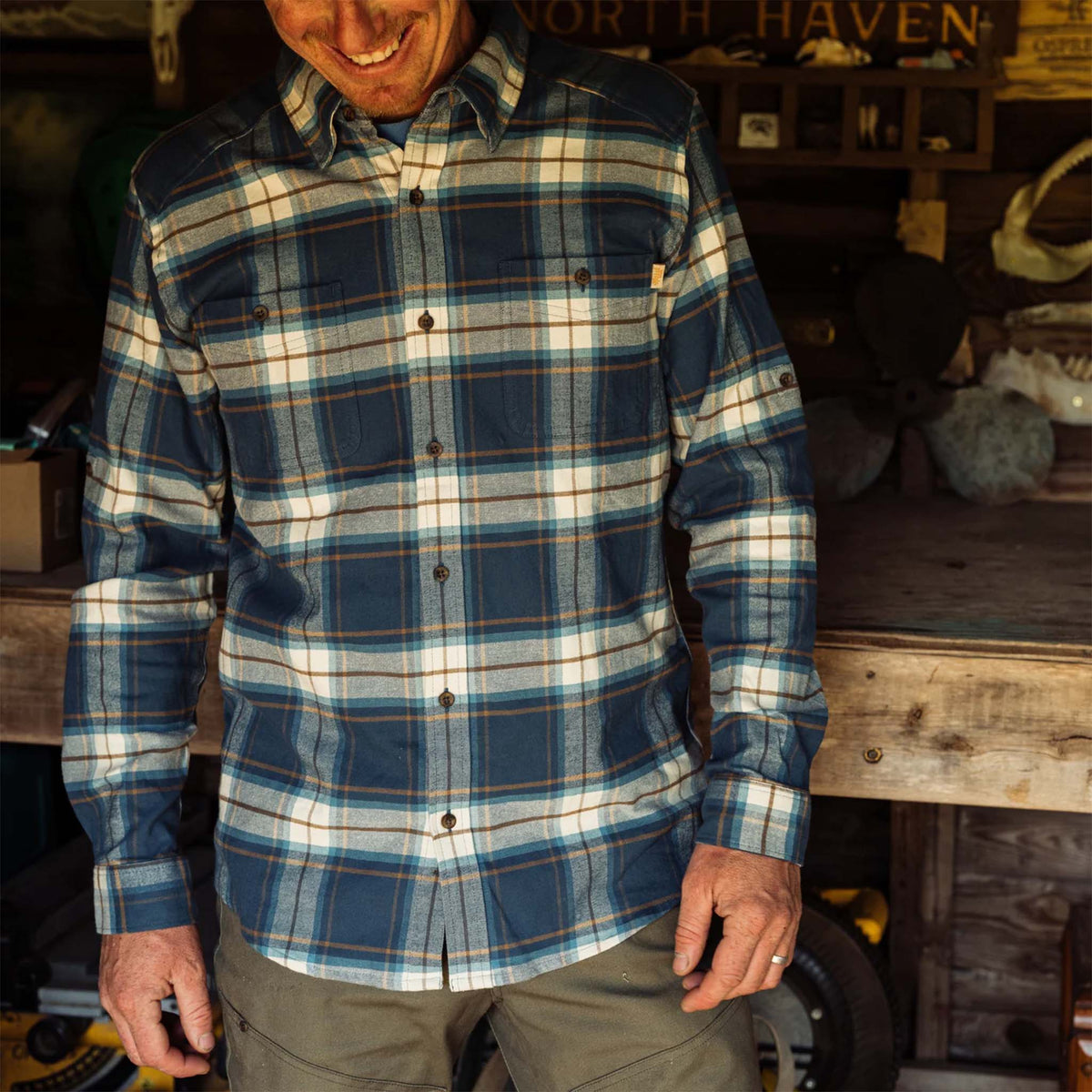 Man wearing a blue plaid shirt in an indoor setting with wooden furniture.