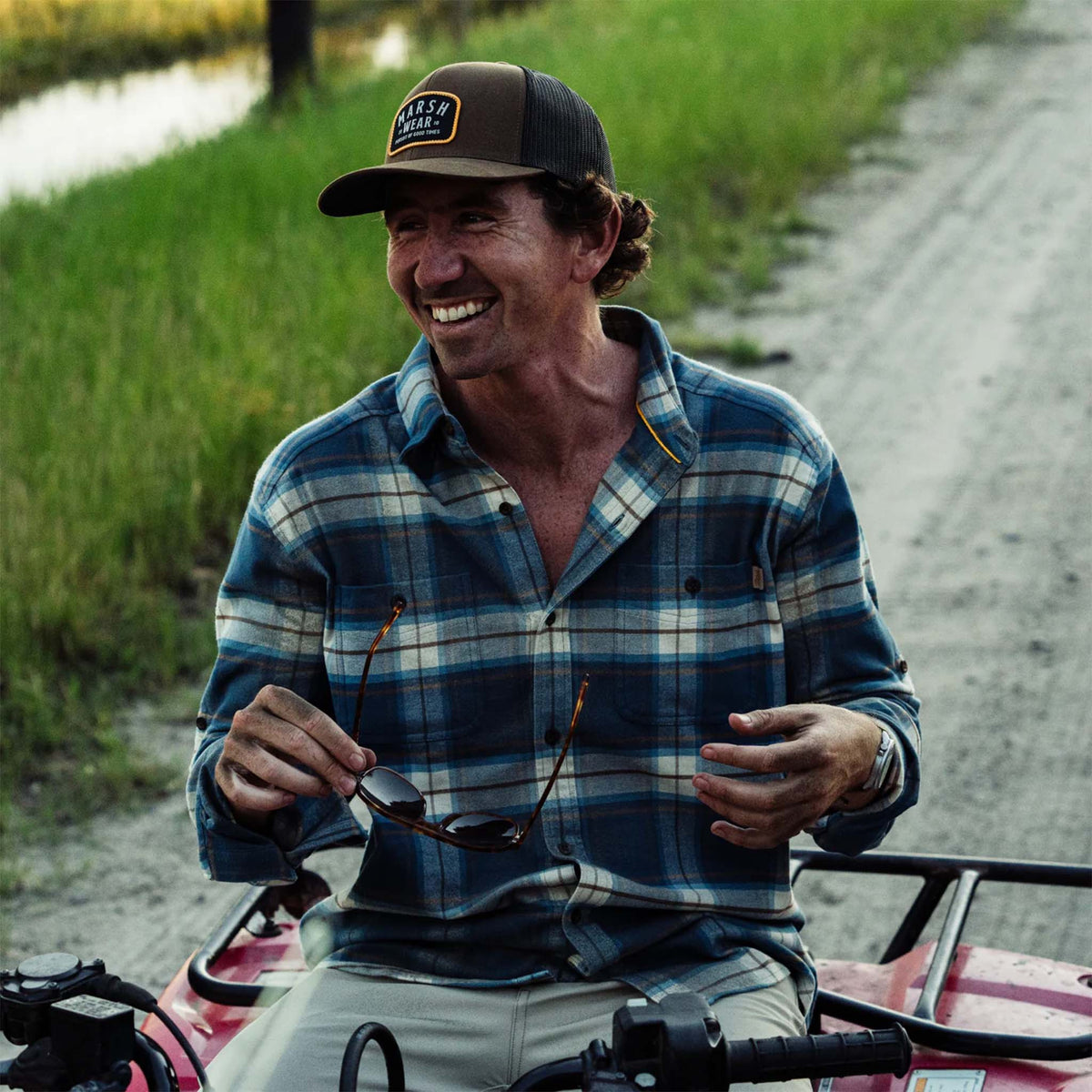 Man sitting on a vehicle outdoors, wearing a plaid shirt and cap.