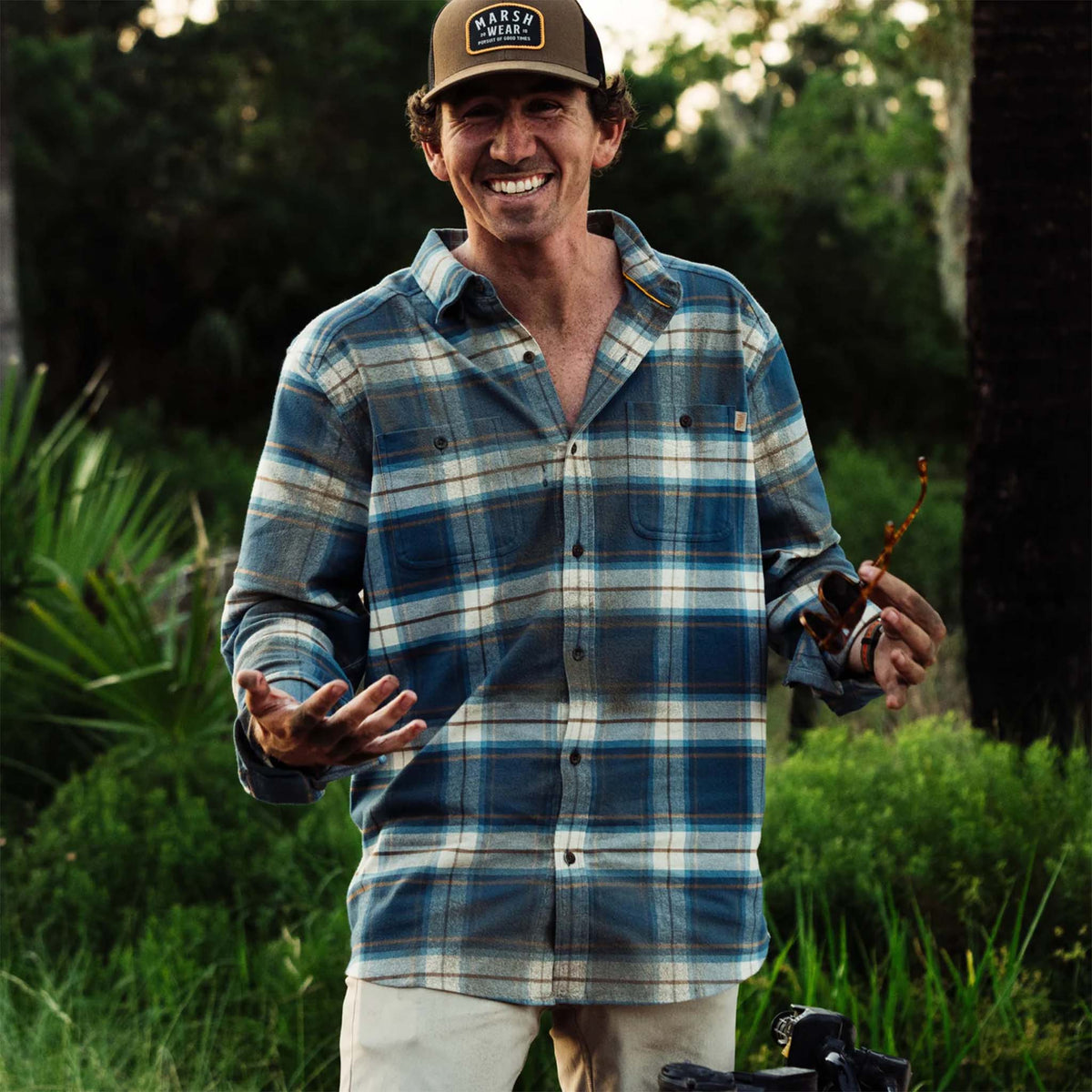 Man wearing a plaid shirt and cap outdoors with greenery in the background