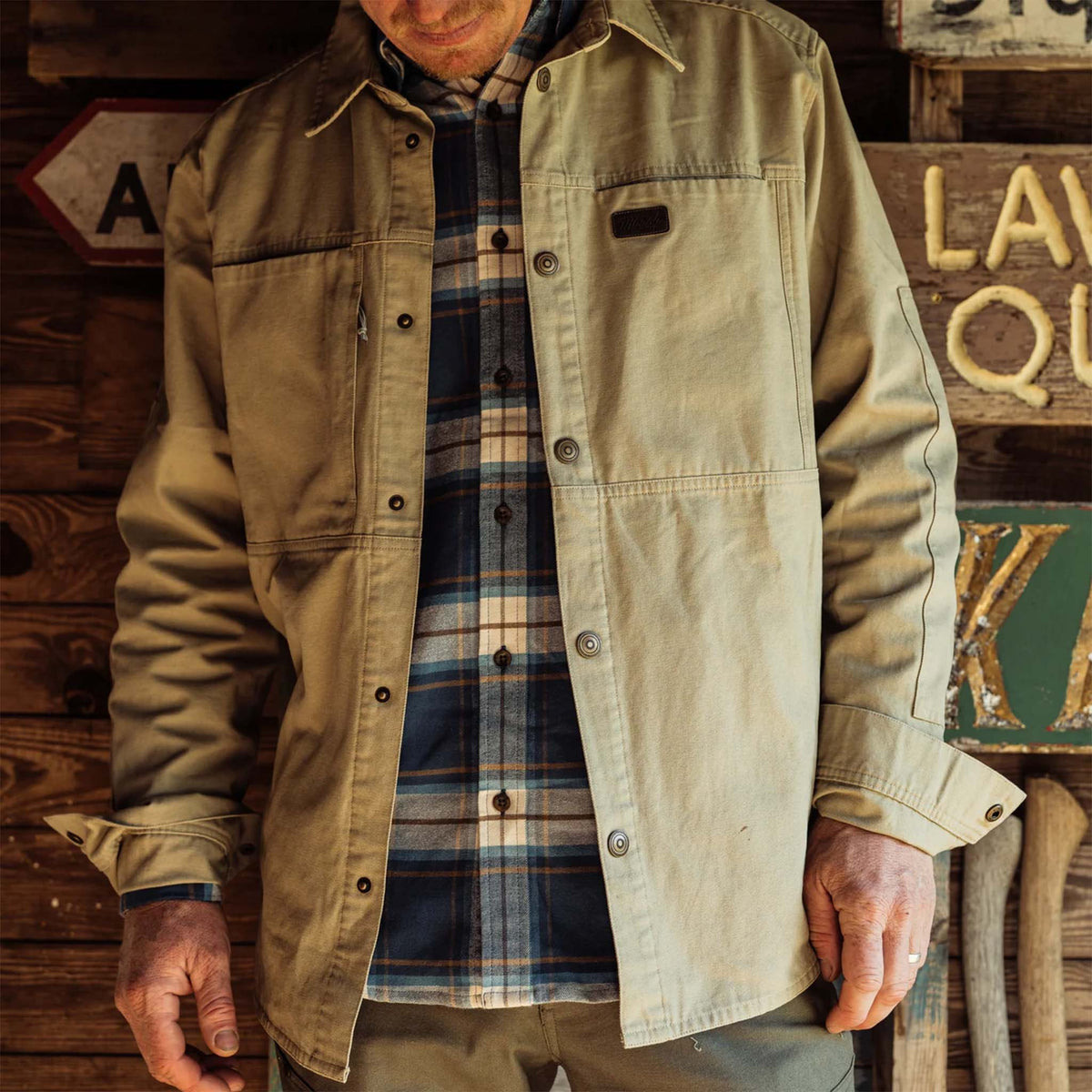 Person wearing a beige jacket over a plaid shirt against a rustic wooden background