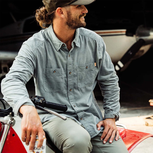 Man sitting on a motorcycle wearing a steel grey shirt and cap, with a blurred background.