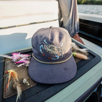 Gray snapback hat with embroidered blue crab graphic and rope detail styled on boat console with fishing lures in a coastal setting.