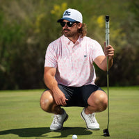 Men’s pink floral short sleeve performance polo styled with black shorts on golf course, paired with cap and sunglasses.