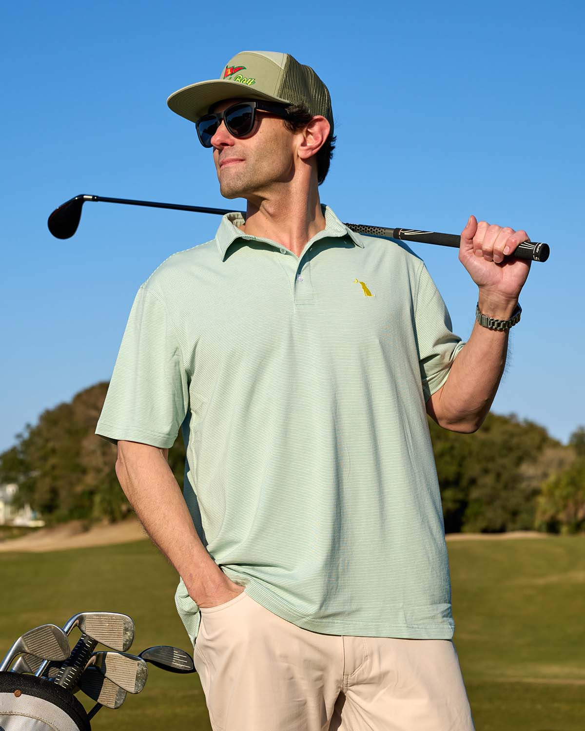 Man wearing light green striped performance polo, khaki shorts, sunglasses, and baseball cap holding a golf club over his shoulder on a sunny golf course.