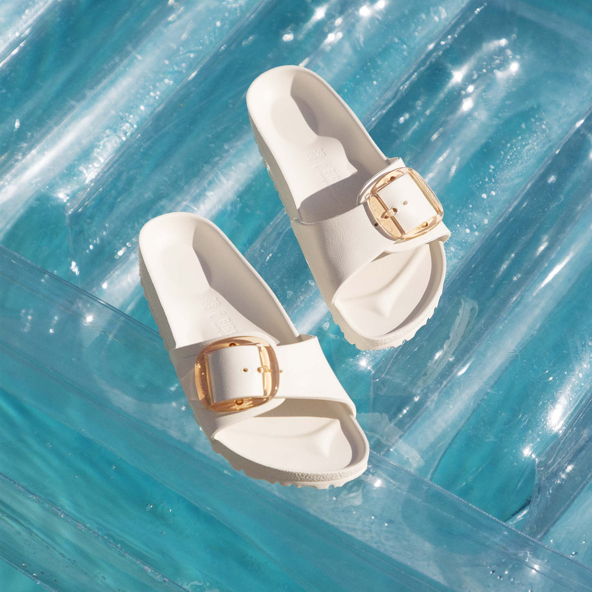 Birkenstock Madrid Big Buckle sandals on a pool float with sun reflection, showing gold buckles and bright white finish.