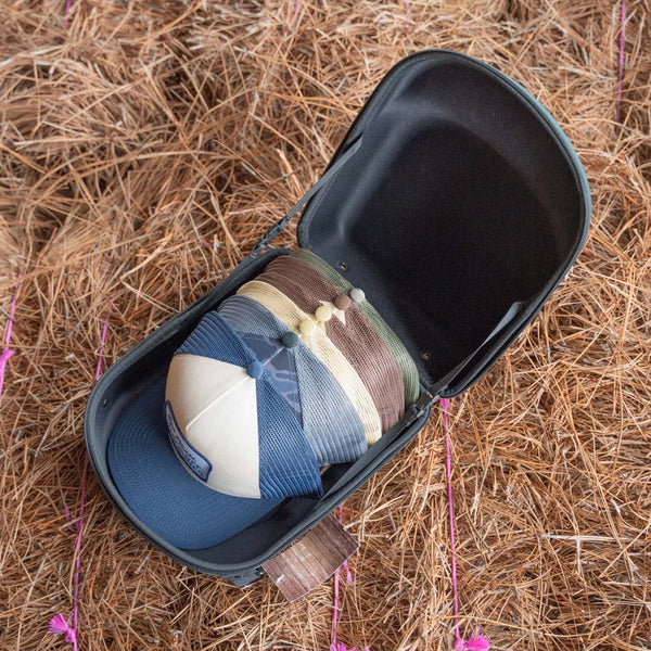 Old South 6 Hat Travel case is green duck pattern.
Open green hard-shell hat case with a duck pattern and black interior, holding 6 stacked trucker hats in various colors, placed on a bed of pine straw.