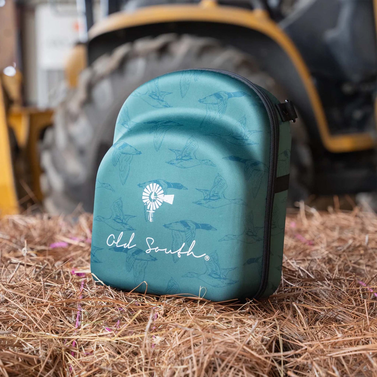 Old South 6 Hat Travel case is green duck pattern.
Closed view of the Old South Duck Pattern Hat Case standing upright on pine straw, featuring a teal green shell with duck illustrations, a white windmill logo, and "Old South" written in cursive. A large tractor wheel is blurred in the background.