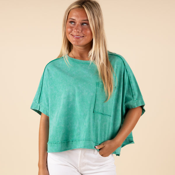  Very J Boxy Pocket Short Sleeve Crop Top, sizes S-L, color green. Woman wearing a boxy green cropped t-shirt with a chest pocket and cuffed short sleeves, styled with white pants.