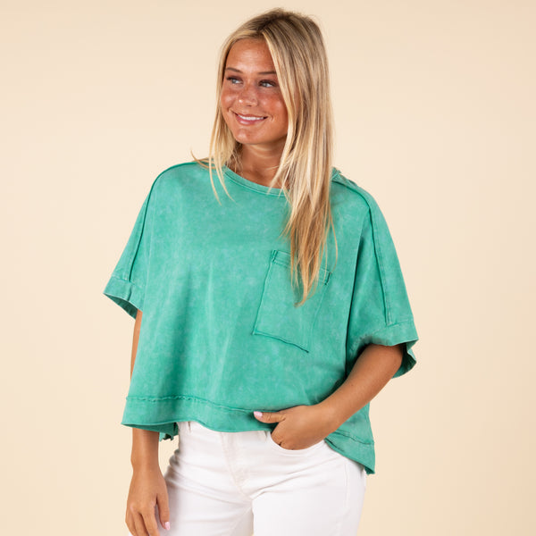  Very J Boxy Pocket Short Sleeve Crop Top, sizes S-L, color green. Woman wearing a boxy green cropped t-shirt with a chest pocket and cuffed short sleeves, styled with white pants.