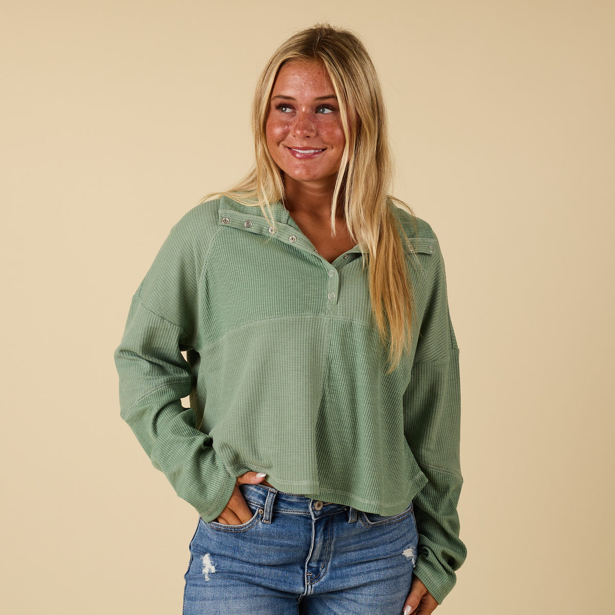 Woman wearing a green long-sleeve shirt and blue jeans against a beige background