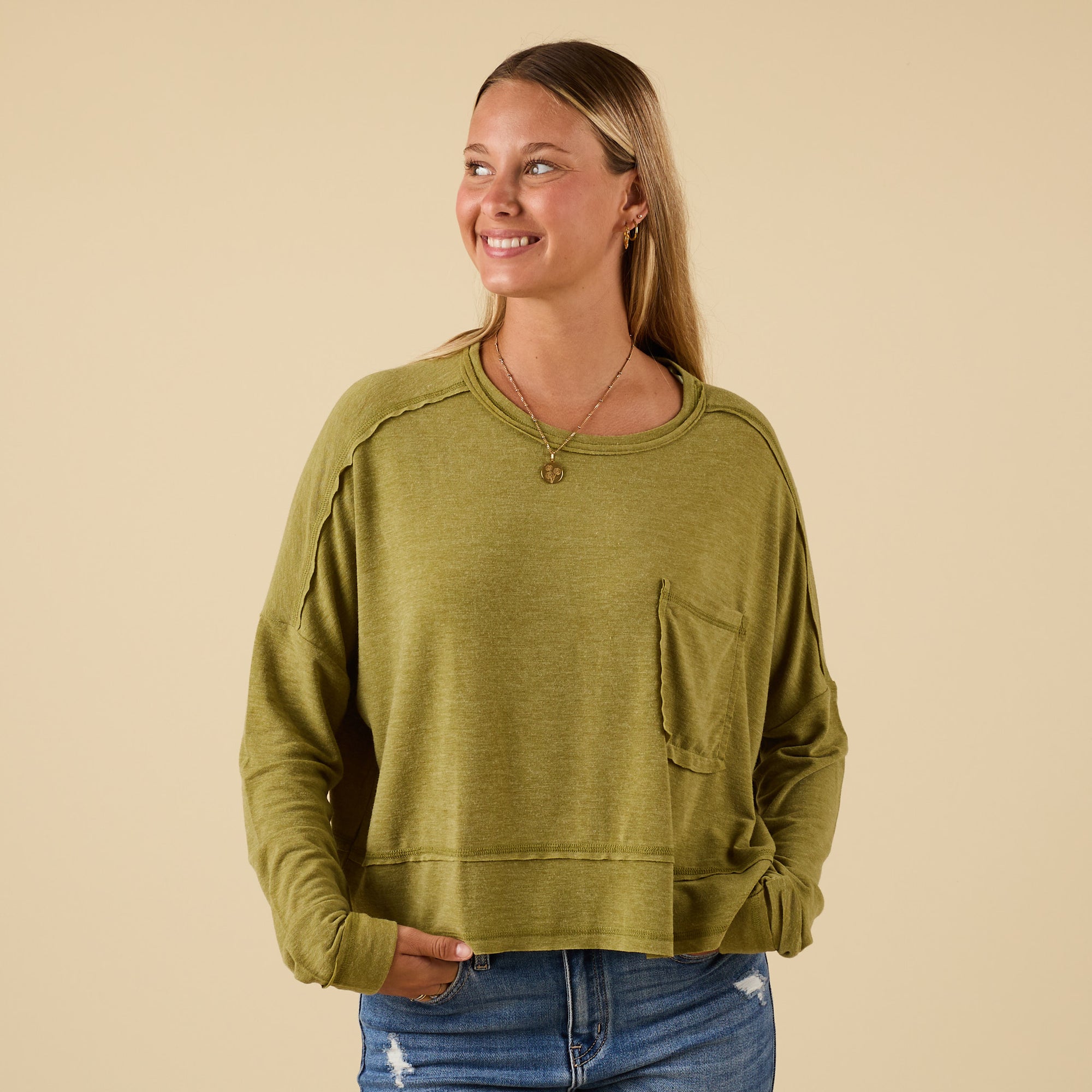 Woman wearing green long sleeve relaxed pocket top with chest pocket and raw seam details, paired with distressed jeans.