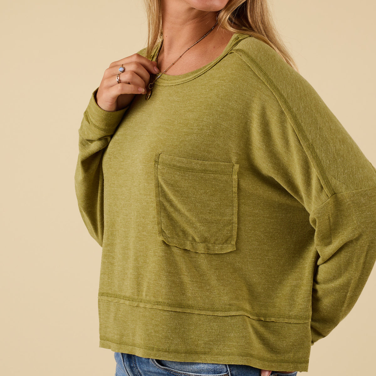 Woman wearing green long sleeve relaxed pocket top with chest pocket, smiling with one hand in pocket, styled with jeans.