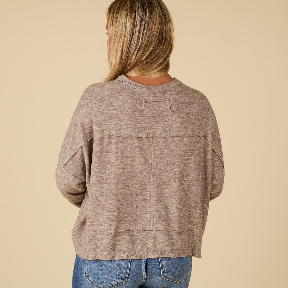Back view of mocha long sleeve relaxed pocket top with raw seam detail and boxy relaxed fit, styled with light wash jeans.