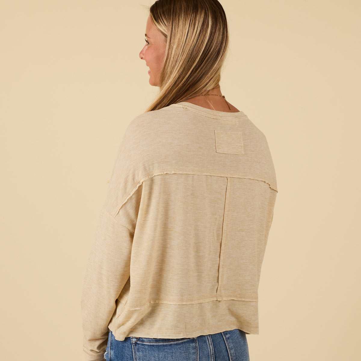 Back view of oatmeal long sleeve relaxed pocket top showing raw seam details across shoulders and center.