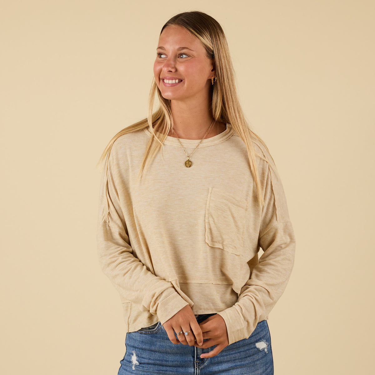 Woman wearing oatmeal long sleeve relaxed pocket top, smiling with hands clasped, styled with blue jeans.