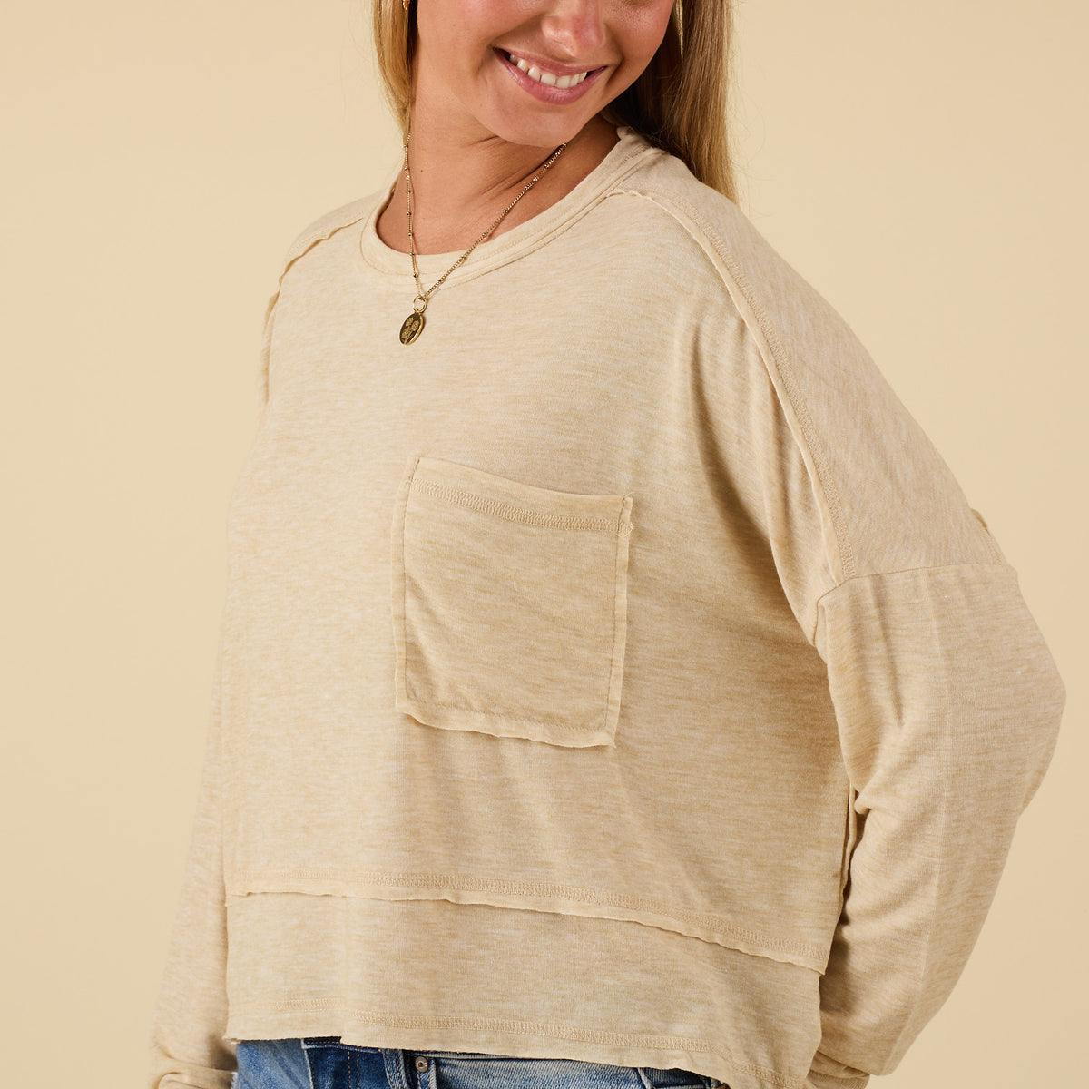 Woman wearing oatmeal long sleeve relaxed pocket top with chest pocket and raw hemline, paired with jeans.