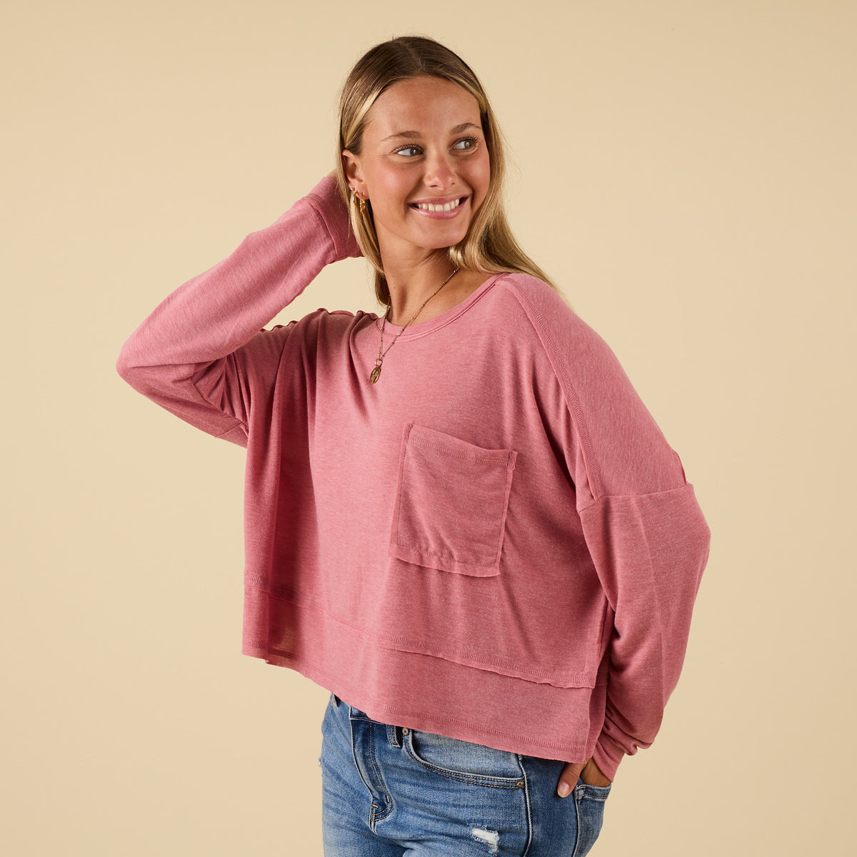 Woman wearing pink long sleeve relaxed pocket top with chest pocket and raw hemline, smiling with hand lifted to hair.