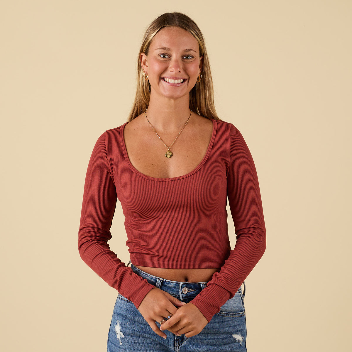 Woman wearing rust ribbed long sleeve scoop neck top with cropped fit and scoop neckline, paired with high-waisted jeans.