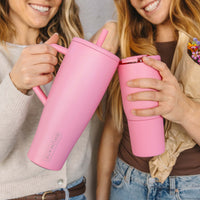 BrüMate pink insulated tumbler and handled mug with straw lids, held by two women clinking drinks in a lifestyle close-up shot.
