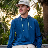 Men’s blue performance hooded long sleeve shirt with contrast drawstrings and small chest logo, worn outdoors in a lifestyle setting with palm trees at sunset.