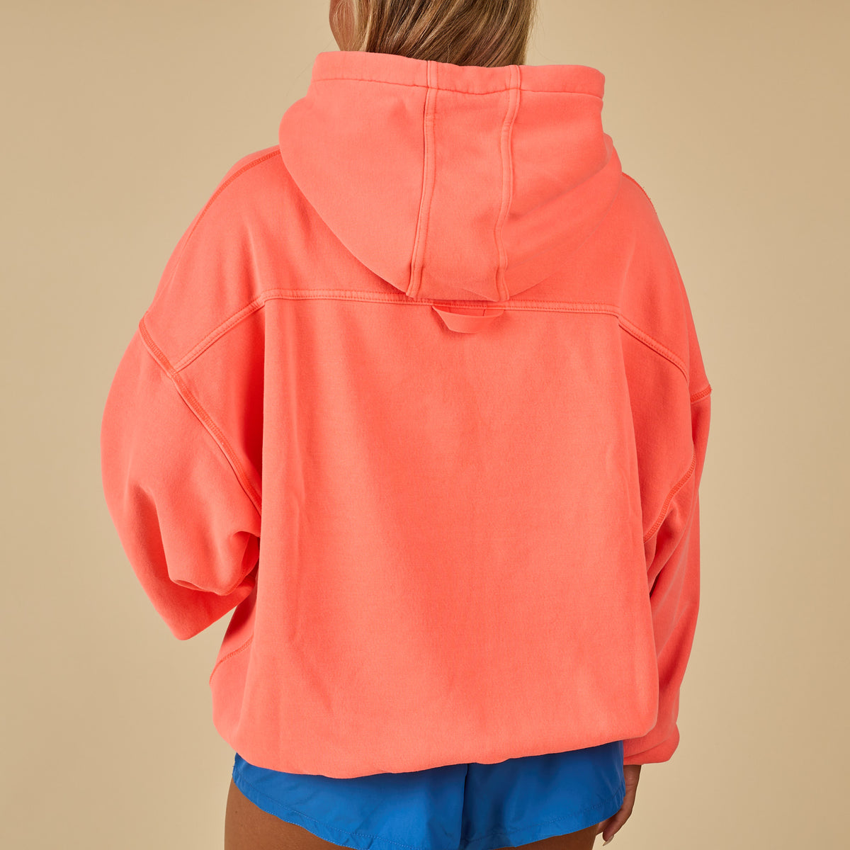 Back view of oversized neon coral hoodie with dropped shoulders and relaxed fit, paired with blue shorts.