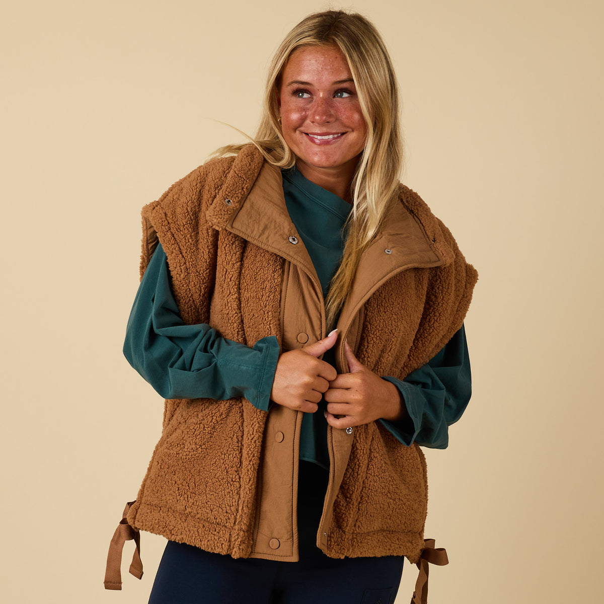 Front view of brown sherpa vest with snap buttons and side ties, worn over a green long sleeve.