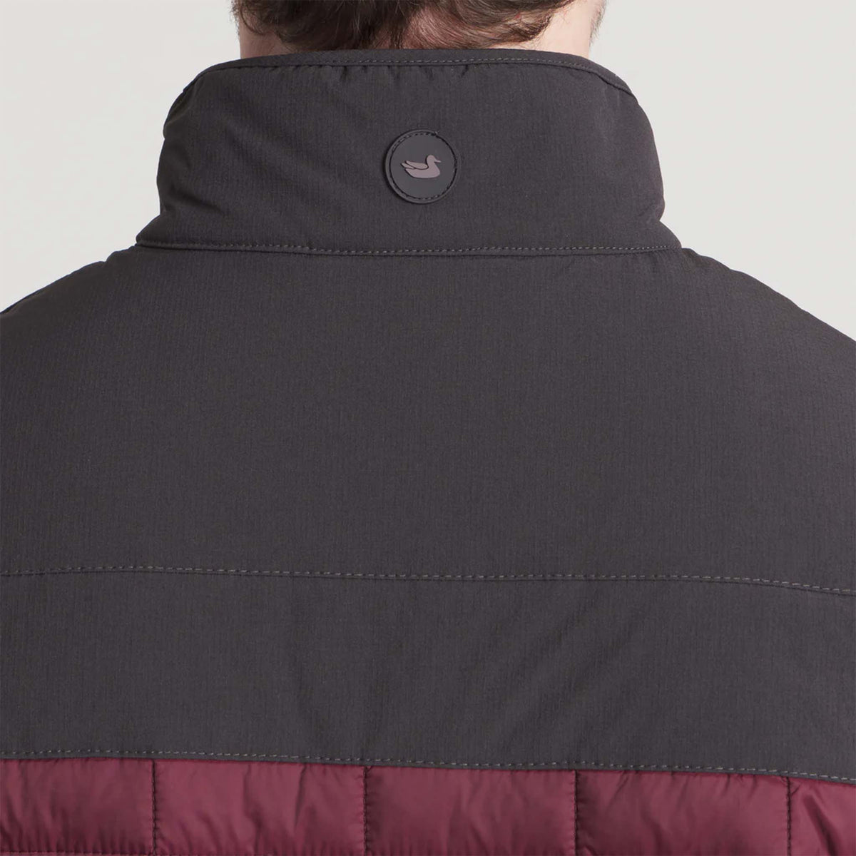 Back view of a person wearing a dark gray and red quilted jacket with a brand logo on a light gray background.