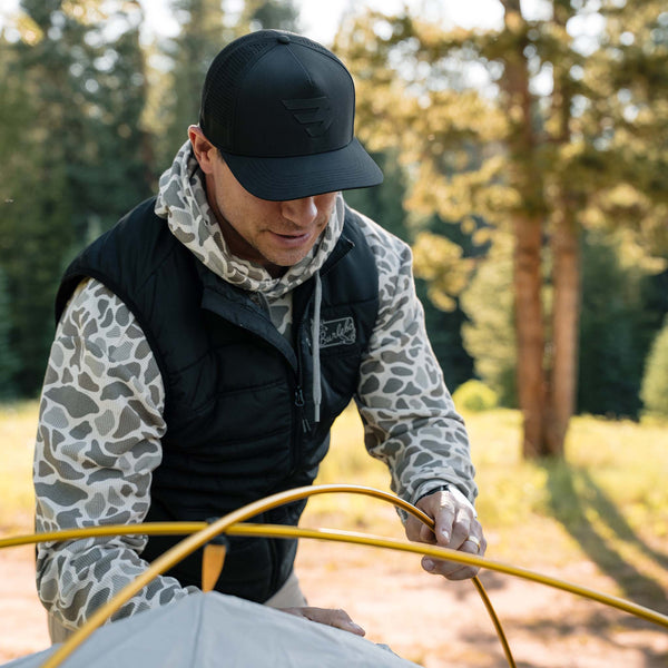 Black performance cap shown in outdoor setting, paired with camo hoodie and black vest.