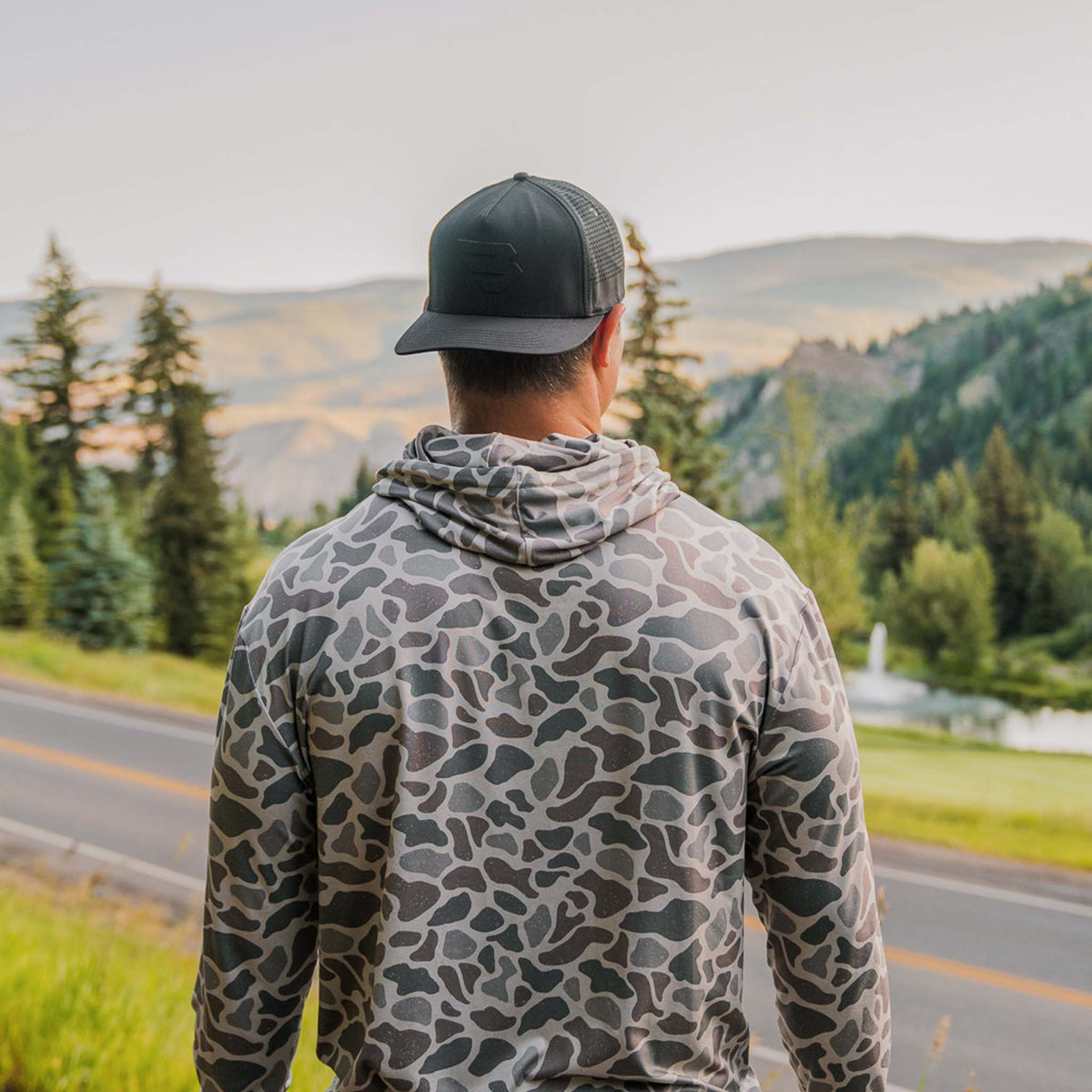 Person wearing a camouflage hoodie and black cap standing on a road with a scenic view of trees and mountains.