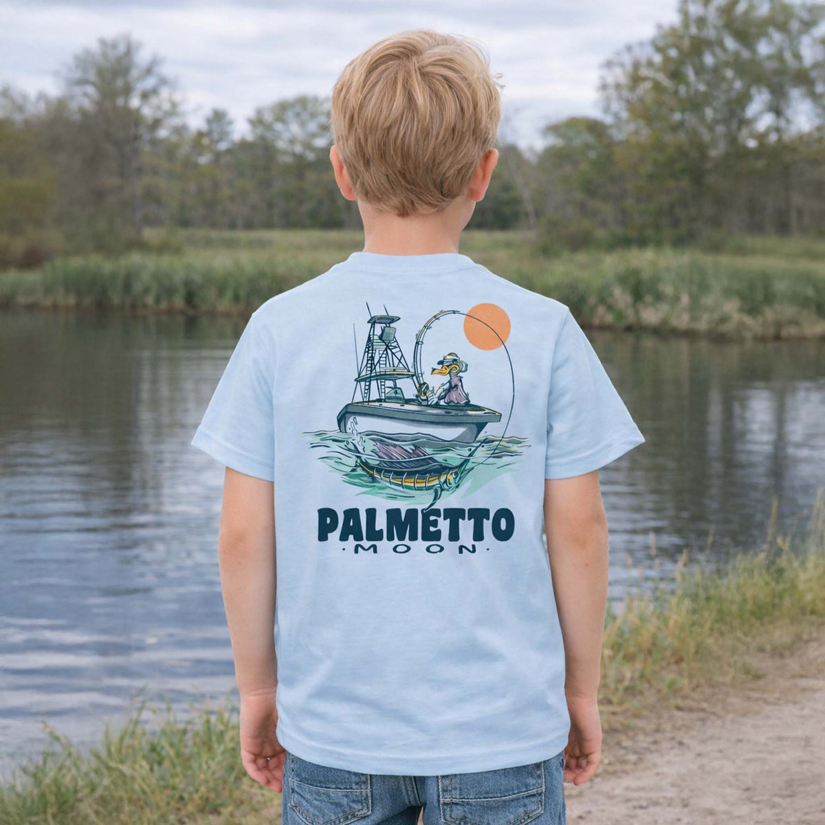 Child wearing baby blue Palmetto Moon youth t-shirt outdoors by a pond showing a back graphic of a duck fishing from a boat with a large fish illustration.