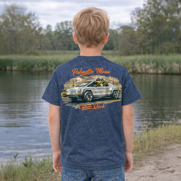 Child wearing heather denim Palmetto Moon youth t-shirt outdoors by a pond showing a back graphic of a duck driving a futuristic truck with “Cyber Duck” text.