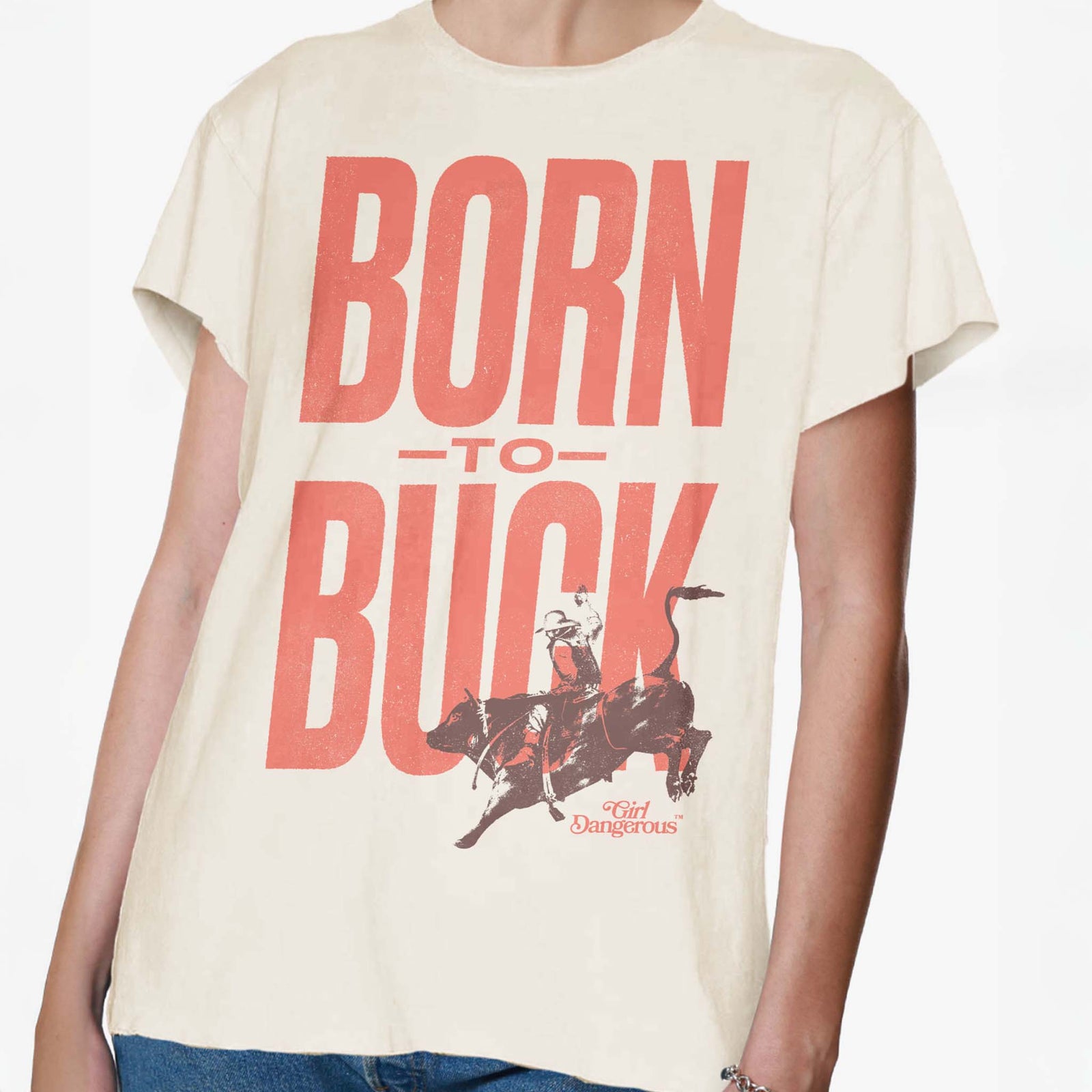 Front view of a natural-colored t-shirt with large red text reading “BORN TO BUCK” and a graphic of a bucking rodeo rider, by Girl Dangerous.