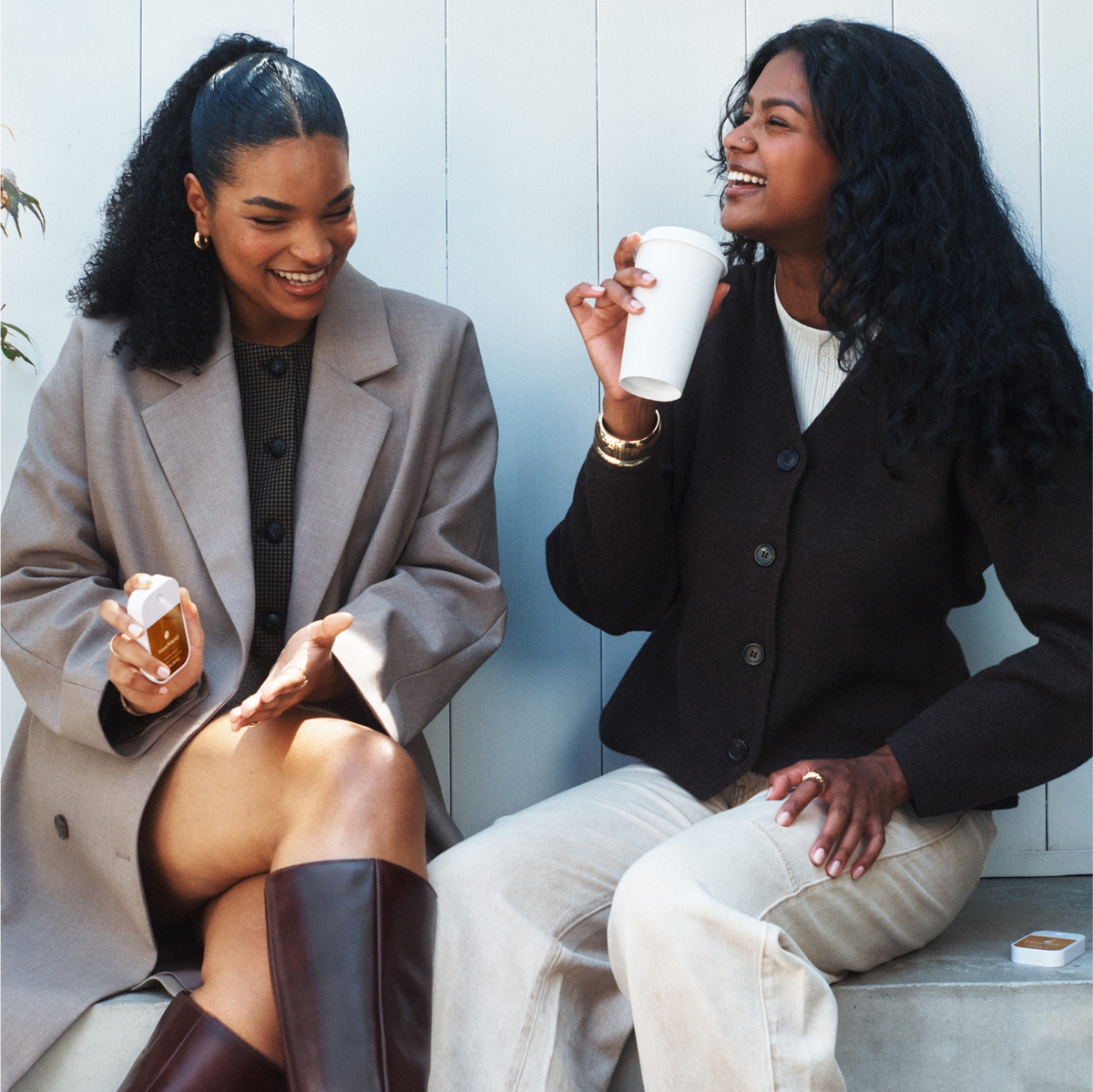 Two women sitting together, one holding a coffee cup and the other a bottle, against a light-colored wall.