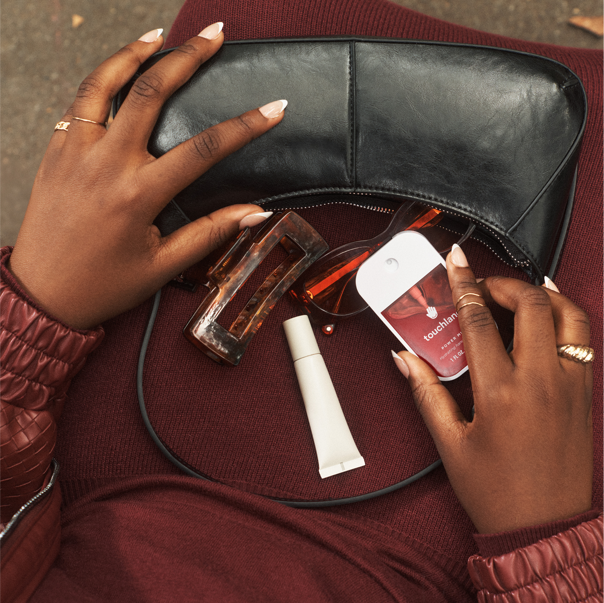 Person opening a black handbag to reveal beauty products including a perfume bottle, cream, and sunglasses.