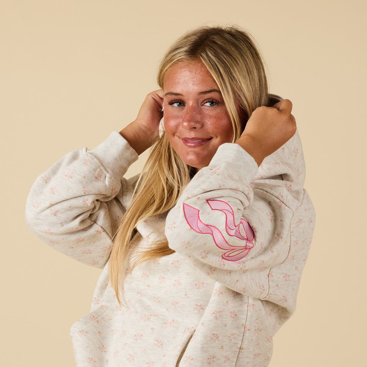 Woman wearing floral hoodie with pink embroidered bow on sleeve, pulling up hood and smiling